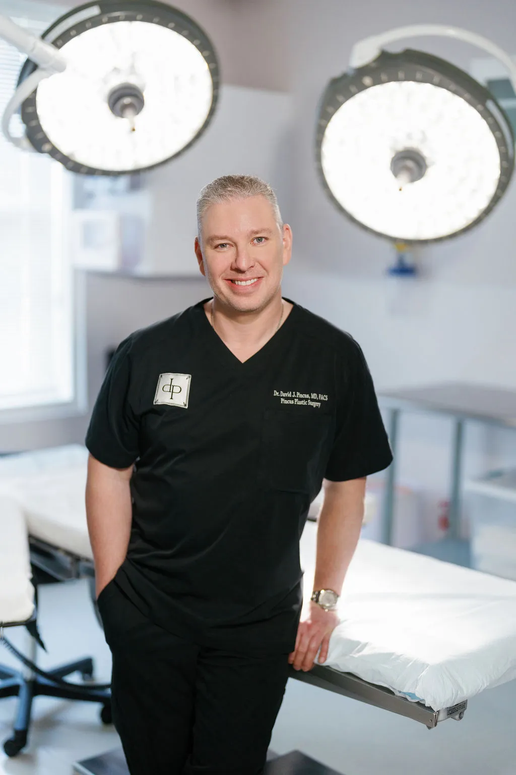 Dr. David J. Pincus, board-certified plastic surgeon, smiling in black scrubs in his operating room.